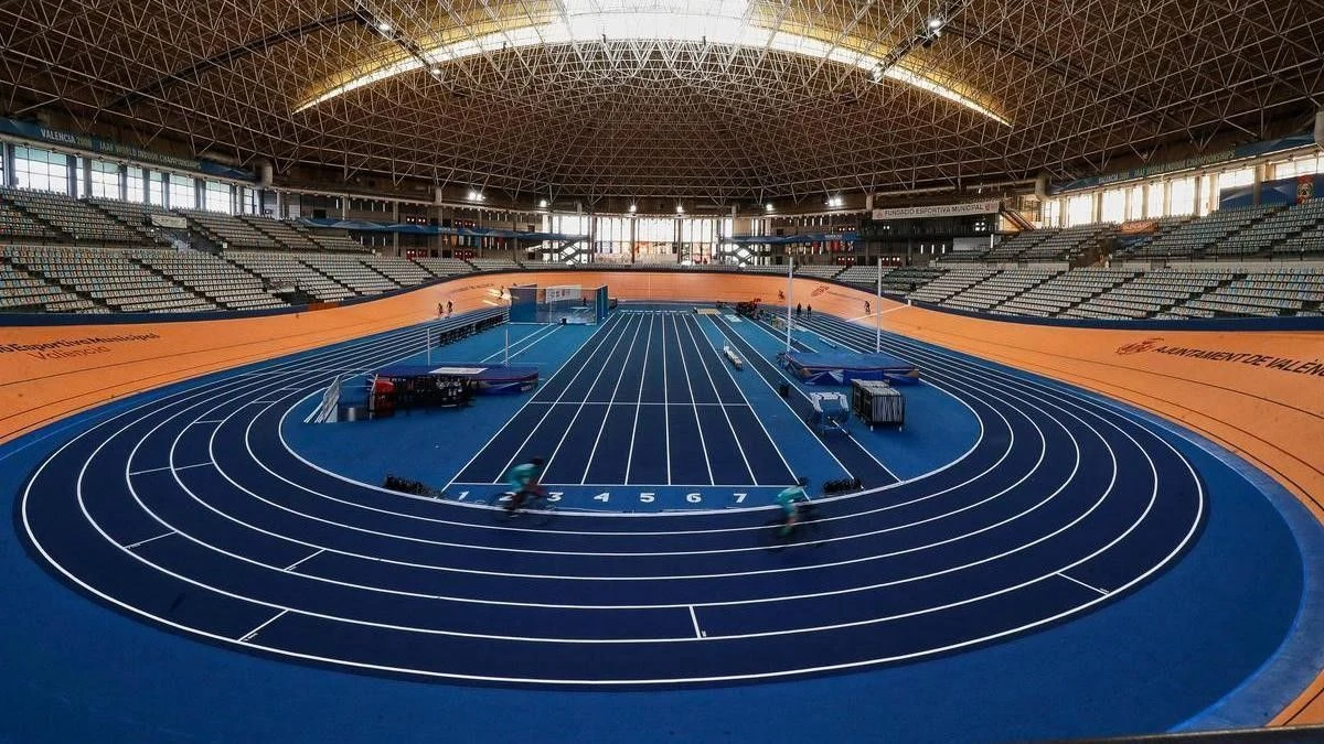Imagen ilustrativa: interior de un velódromo cubierto con pista de atletismo y gradas vacías, iluminado por luz lateral.