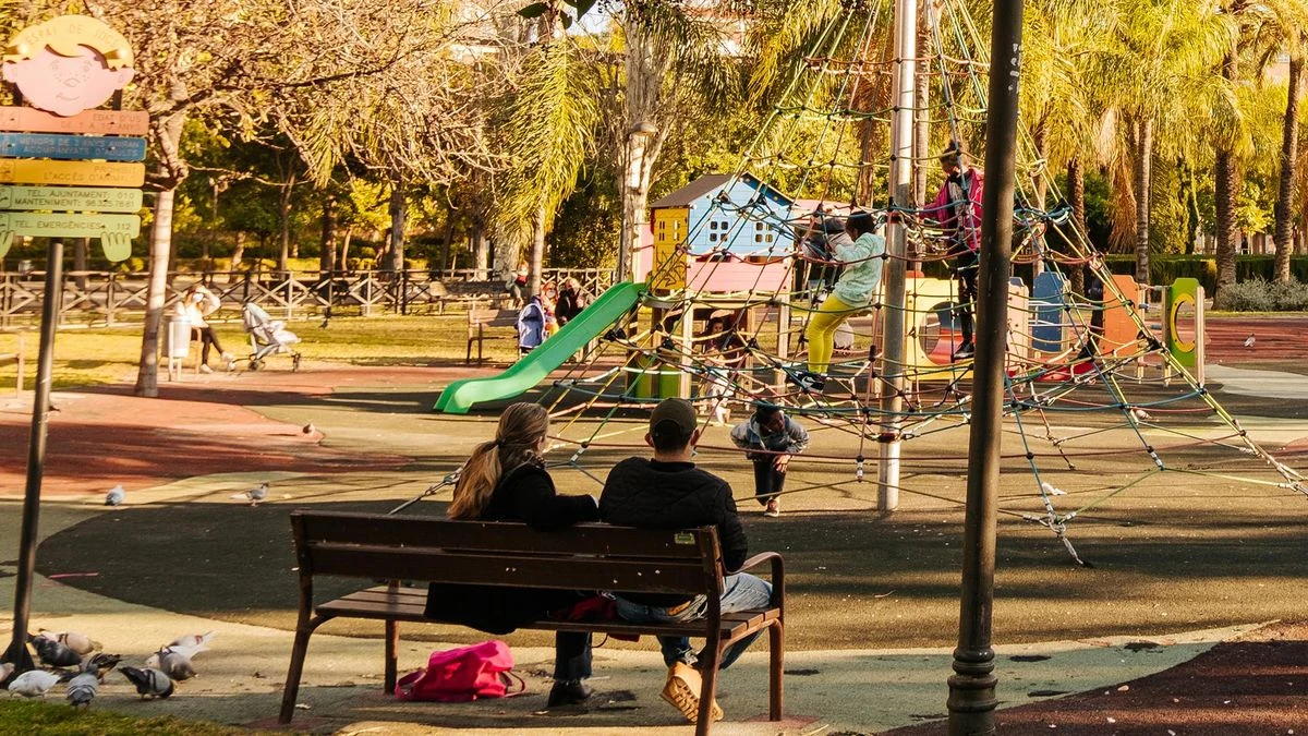 Imagen ilustrativa: un parque infantil con columpios y toboganes bajo la sombra de árboles, con una cámara fotográfica en primer plano