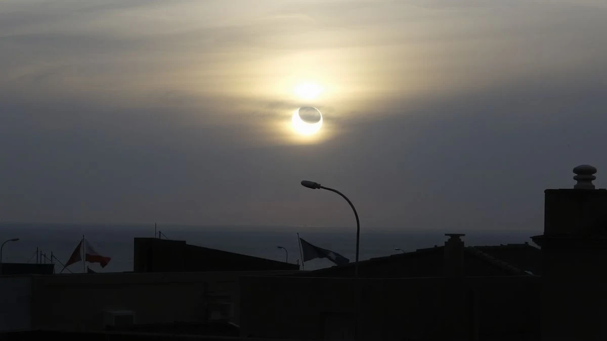 Imagen ilustrativa: un eclipse solar parcial sobre el horizonte marino de una localidad costera, con siluetas de edificios y algunas personas observando de espaldas.